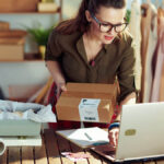 Business owner holding a package and checking her laptop.