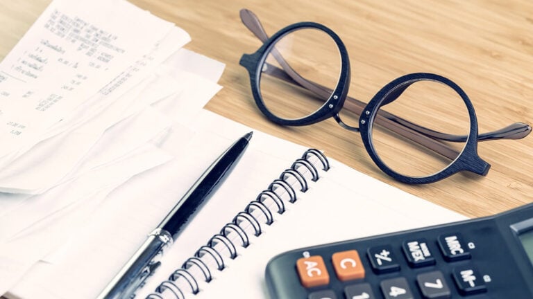 Glasses, notebook, calculator, pen, and receipts on a desk.