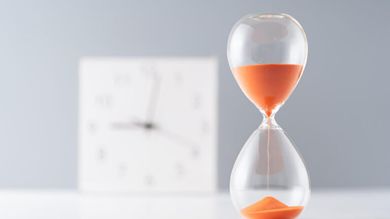 Hour glass with orange sand. Clock in background.