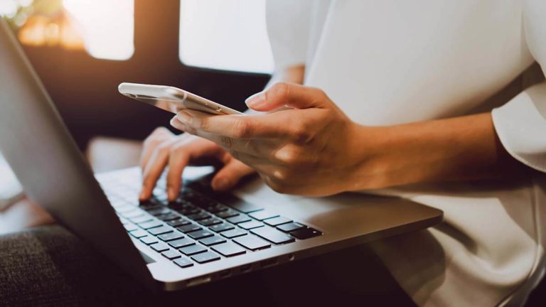 Hands holding a cell phone and using a laptop.