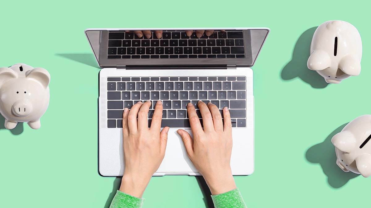 Hands on a laptop with piggy banks on the table.