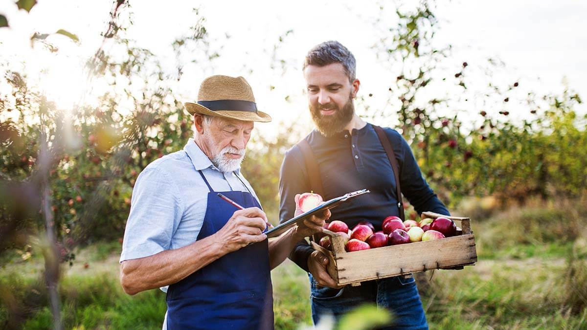 Man holding a bushel of apples and talking with a man taking inventory outside.
