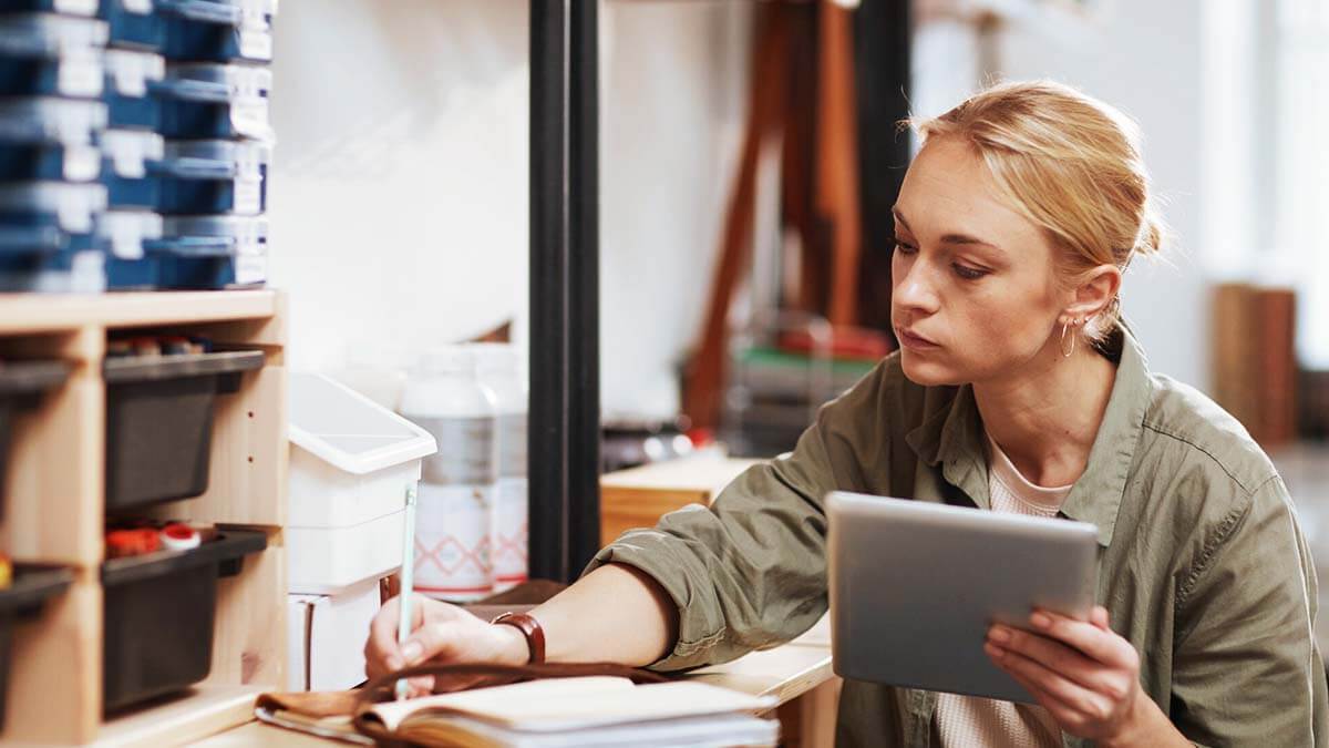 Woman holding a tablet and writing in a notebook.