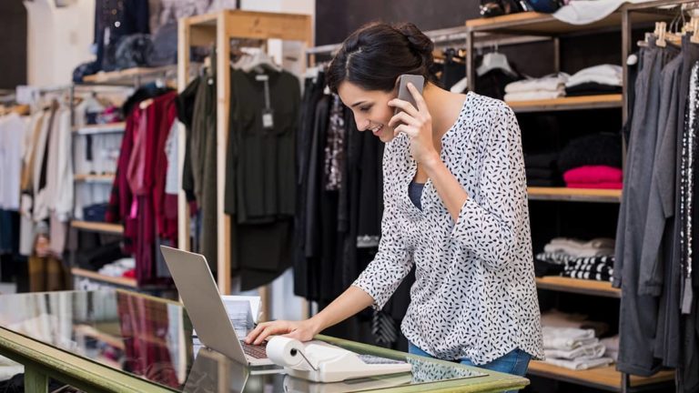 woman on phone using laptop in a clothing store, creating a sales journal entry