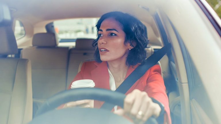 woman in orange blazer driving and holding a cup of coffee while looking in the rearview mirror
