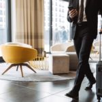 businessman wearing formal black suit holding smartphone and walking with suitcase in hotel lobby
