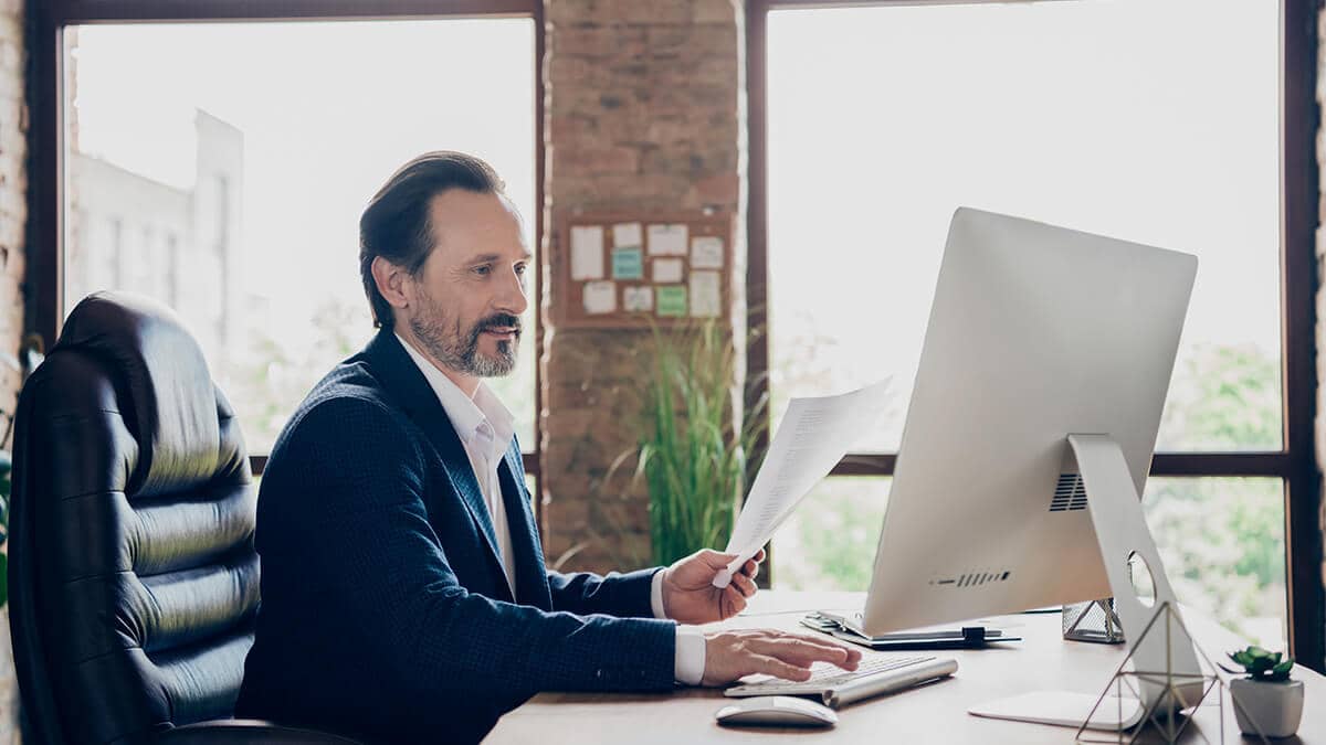 Man holding up a piece of paper while using a desktop computer.