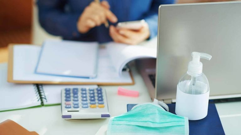 Desk with a calculator, hand sanitizer, laptop, and paperwork on it.