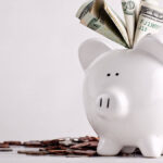 White piggy bank with bills sticking out and coins on the ground.