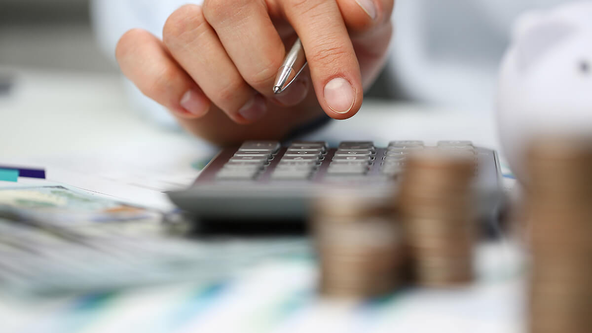 Hand typing on a calculator with stacks of coins.