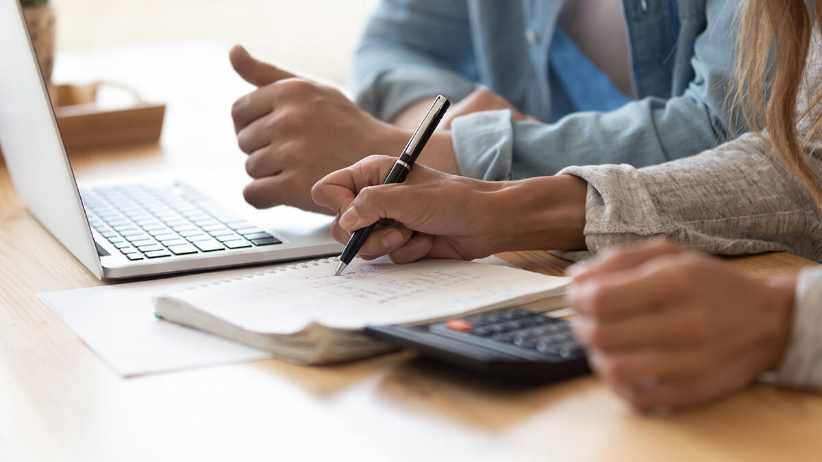 Two accountants writing on a piece of paper and using a laptop and calculator.