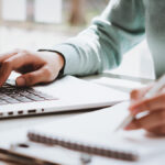 Business owner writing on spiral notepad and using laptop.