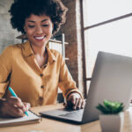 Smiling woman writing in a notebook and using a laptop.