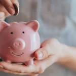 Woman putting a coin into a pink piggy bank, representing Trump Accounts.