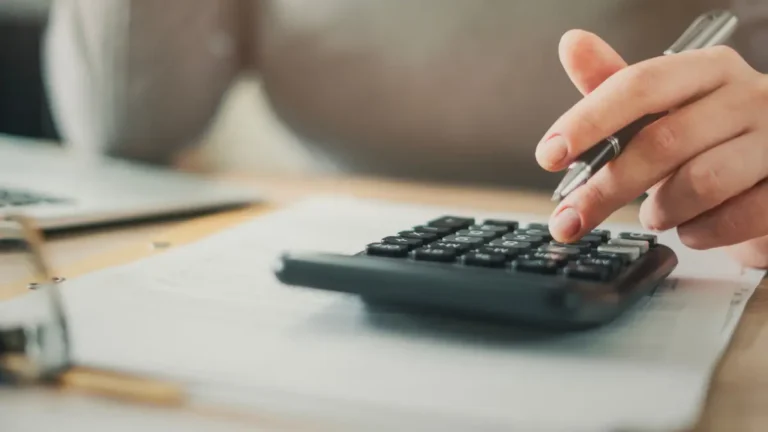 Close-up of a person holding a pen and using a desk calculator to calculate payroll deductions.